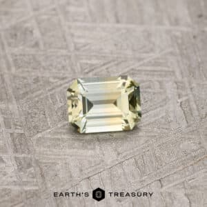 A 0.79-carat, yellow-pale aqua particolored Montana sapphire with an emerald cut rests on a textured gray surface. The gemstone features precise, symmetrical facets that enhance its clarity and brilliance. The text "EARTH'S TREASURY" is printed below the stone.