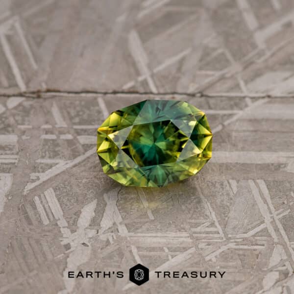 A faceted, oval-shaped gemstone featuring a gradient from yellow to green rests on a textured surface. The background has an intricate, industrial design. Below the gemstone, the text reads "2.74-Carat Lime Green Australian Sapphire.