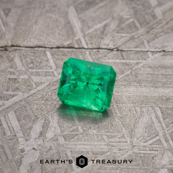 A 2.13-carat bright green emerald, rectangular-cut, rests on a textured, grayish surface. The emerald showcases a rich, vibrant color with visible facets and internal inclusions that enhance its character. The background features the text "EARTH'S TREASURY" at the bottom center.