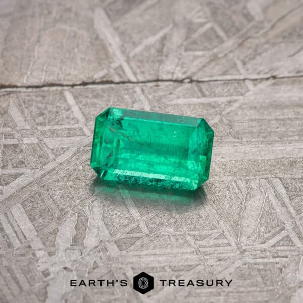 A vibrant 2.13-carat bright green emerald gemstone cut into a rectangular shape rests on a textured, patterned surface. The image features a logo at the bottom that reads "Earth's Treasury" with a small geometric design beside it.