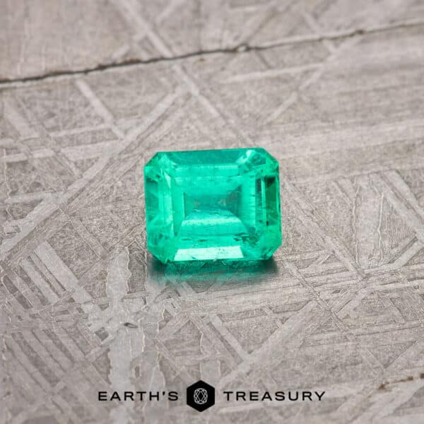 A 2.13-carat, bright green, emerald-cut gemstone is placed on a textured grey surface. "Earth's Treasury" is printed below the gemstone in black text.