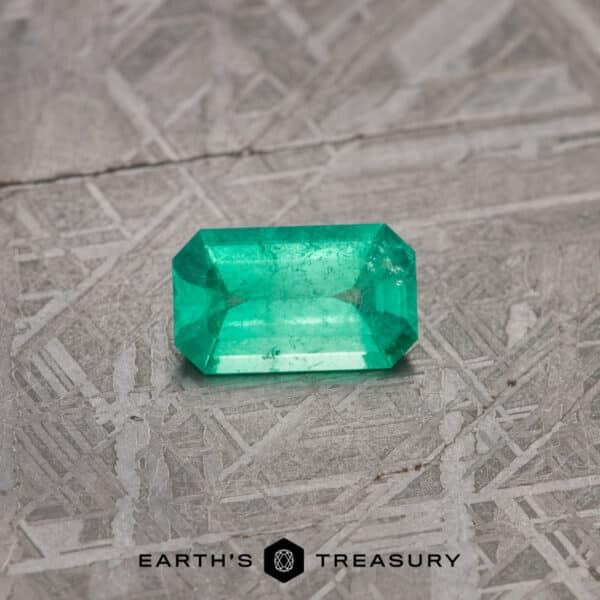 A vibrant, rectangular-cut 2.13-carat bright green emerald is showcased on a textured gray surface. The imagery features the brand logo "EARTH'S TREASURY" at the bottom. The emerald displays a rich green hue with visible internal characteristics typical of natural stones.