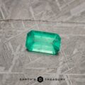 A 2.13-carat bright green emerald with a faceted cut rests on a textured gray surface. "EARTH'S TREASURY" is written at the bottom of the image with a geometric logo between the words.