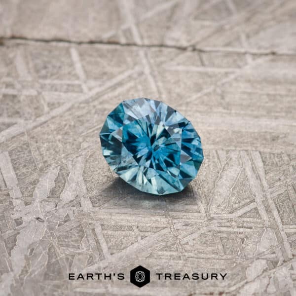 A round, 1.22-carat medium blue Montana sapphire with intricate facets is displayed on a textured gray surface. The text "EARTH'S TREASURY" is printed below the gemstone.