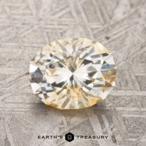 A 1.93-carat, faceted round yellow sapphire from Montana sits delicately on a textured gray surface, accompanied by the brand name "Earth's Treasury" at the bottom.