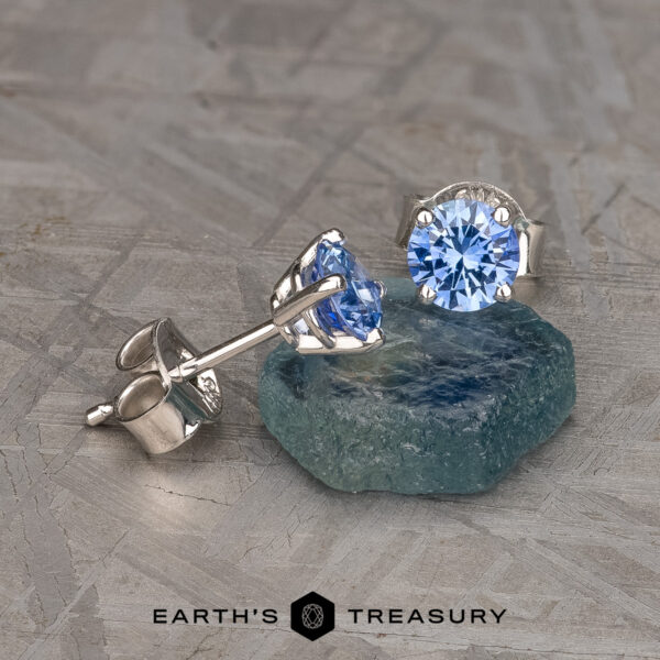 Two Medium Blue Sapphire Earrings, 3.5-5.0 mm (Heated), each set in silver prong settings, are artistically arranged on a textured gray surface. One earring is laid flat while the other stands upright, supported by a rough teal stone. The "Earth's Treasury" logo is at the bottom center.