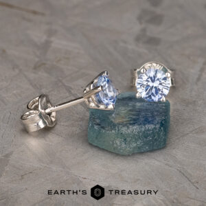 A pair of Light Blue Sapphire Earrings, 3.5-5.0 mm (Heated), adorn elegant faceted blue stones set in silver posts, displayed beautifully on a rough aquamarine crystal. The earrings shine prominently against a textured gray surface, with the text "EARTH'S TREASURY" and a hexagonal logo at the bottom.