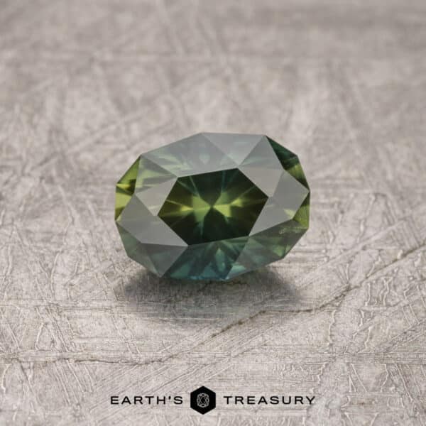 A 1.34-carat Deep Green-Lime Particolored Australian Sapphire with a faceted cut, showcasing vibrant green and blue hues, is displayed on a textured gray surface. The words "Earth's Treasury" are visible at the bottom.