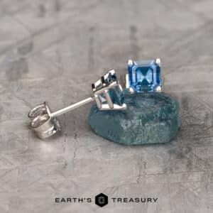 A pair of Montana Sapphire Earrings- Asscher, 4.5mm (Heated) in silver is displayed on a textured surface. One earring is positioned upright, showcasing the square-cut blue gemstone, while the other is laid on its side. The brand name "EARTH'S TREASURY" is visible at the bottom of the image.