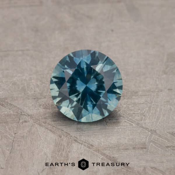 A 2.14-carat robin's egg blue Montana sapphire (heated) is displayed on a textured gray surface. The gemstone has a brilliant cut, showcasing various facets that reflect light beautifully. Below the gemstone, the text "Earth's Treasury" is visible.