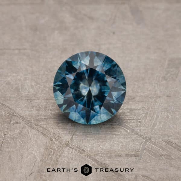 A 2.14-carat Robin's Egg Blue Montana sapphire gemstone, with intricate facets, is displayed on a textured, light gray surface. The sapphire is positioned above the logo and the text "EARTH'S TREASURY" in black font.