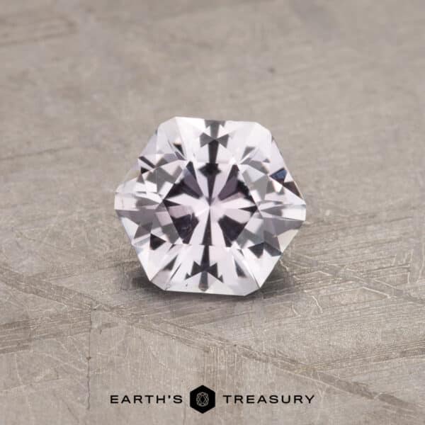 A faceted hexagonal 0.91-carat near colorless Montana sapphire with a brilliant cut, displaying clear and sparkling clarity, is placed on a textured gray surface. The text "EARTH'S TREASURY" is centered below the gemstone.