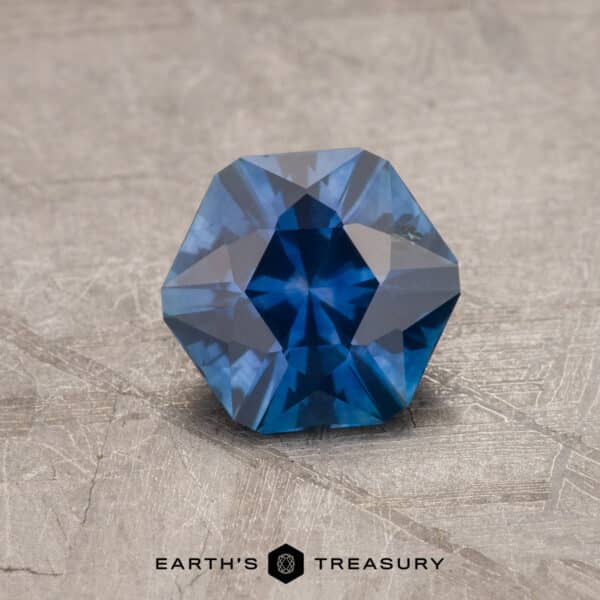 A close-up image of a faceted 2.14-carat Robin's egg blue Montana sapphire, shaped in a hexagonal pattern with intricate cuts reflecting light. The gem is placed on a textured gray surface, with the text "Earth's Treasury" written below in black font.