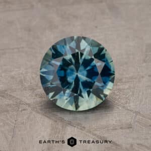 A close-up of a round, faceted 2.14-Carat Robin's Egg Blue Montana Sapphire (Heated) showcasing its exquisite blue and green hues. It is placed on a textured, gray surface with the text "Earth's Treasury" visible beneath the gemstone.