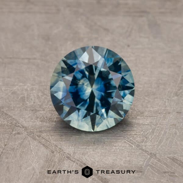 A close-up of a round-cut, robin's egg blue Montana sapphire gemstone weighing 2.14 carats, placed on a textured, neutral-colored surface. The gemstone features a mix of light and dark hues with intricate facets reflecting light. The logo "Earth's Treasury" is visible at the bottom of the image.