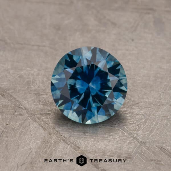 A close-up image of a round, 2.14-carat Robin's Egg Blue Montana Sapphire with intricate facets reflecting light, placed on a textured, gray surface. The text "EARTH'S TREASURY" and a small hexagonal logo are positioned at the bottom of the image.