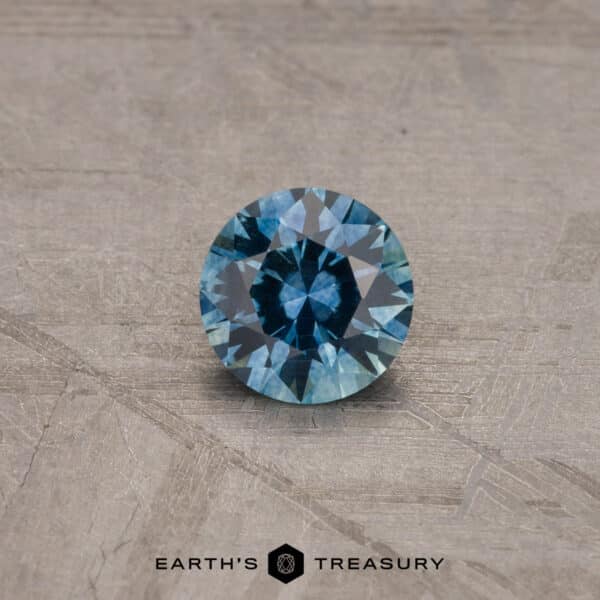 A round, robin's egg blue 2.14-carat Montana sapphire gemstone is displayed against a textured gray background. The gem's cut reveals intricate facets, and the text "Earth's Treasury" is visible at the bottom of the image.