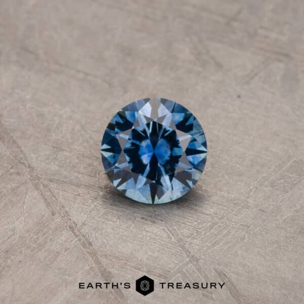 A 2.14-carat Robin's Egg Blue Montana Sapphire (Heated) displayed on a textured, gray surface. The stone's round brilliant-cut facets reflect light, showcasing its vibrant hue. Below the gemstone, the text "Earth's Treasury" is visible, along with a geometric logo.