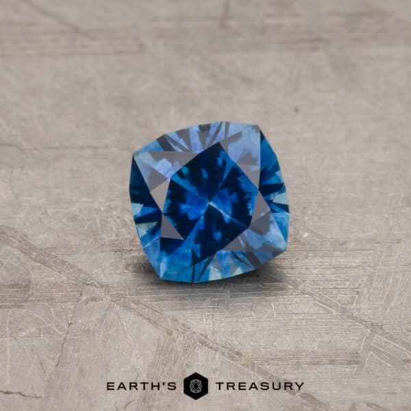 A close-up image showcases a 2.14-carat Robin's Egg Blue Montana Sapphire (Heated) on a textured, light-colored surface. The cushion-shaped gemstone exhibits various shades of blue, and the words "EARTH'S TREASURY" are visible in the bottom center.