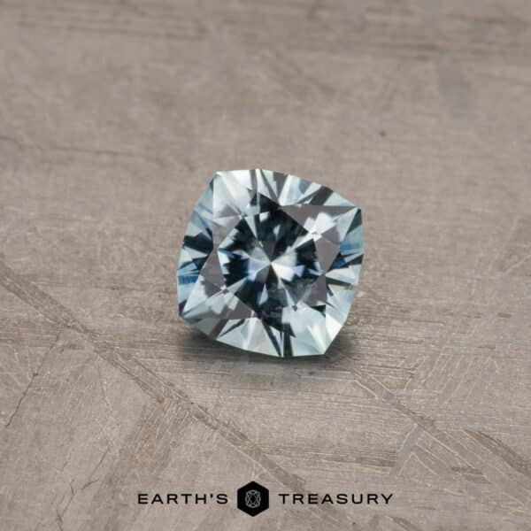 A 2.14-carat Robin's Egg Blue Montana Sapphire (heated) rests on a textured, gray background. The gemstone displays a brilliant mix of light blue hues and intricate facets, capturing light beautifully. The logo "Earth's Treasury" is positioned below the sapphire.
