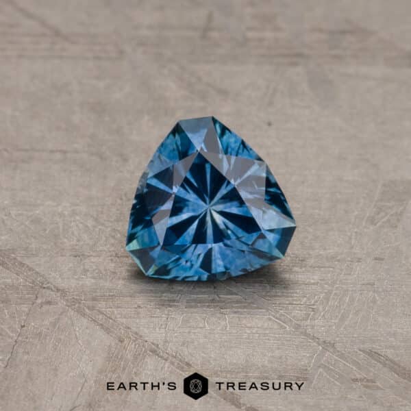 A brilliant, blue trillion-cut gemstone with intricate facets sits on a textured gray background. The gem is likely a 2.14-Carat Robin's Egg Blue Montana Sapphire (Heated) due to its vivid color and sparkle. The text "EARTH'S TREASURY" is visible at the bottom center.