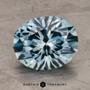 A 2.14-carat Robin's Egg Blue Montana Sapphire (heated) is centered on a textured gray surface. The gem exhibits a vivid sparkle and intricate cuts, showcasing various shades of blue. Below the gemstone, "Earth's Treasury" is printed in black text.