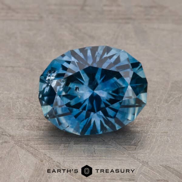 A 2.14-carat Robin's Egg Blue Montana Sapphire (heated) sits on a textured gray surface, exhibiting intricate internal patterns and brilliant facets that reflect light. The text "EARTH'S TREASURY" is seen at the bottom of the image.