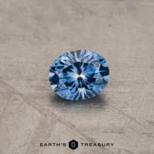 A brilliant blue gemstone, the 2.14-Carat Robin's Egg Blue Montana Sapphire (Heated), sits on a textured, beige surface. The gem is faceted and catches the light, showcasing its sparkling, vivid blue color. The words "EARTH'S TREASURY" are visible at the bottom of the image.