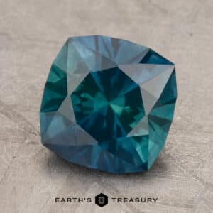 A 2.14-carat Robin's Egg Blue Montana Sapphire, with heated treatment, displays intricate facets reflecting light as it rests on a textured gray surface. The words "EARTH'S TREASURY" are visible at the bottom of the image.