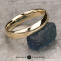 A polished gold The Classic Band, Comfort Fit ring rests on a piece of raw blue stone. The interior of the ring features an engraved logo and the letters "MAK." The background is a textured, stone-like surface. The text "EARTH'S TREASURY" appears at the bottom center of the image.