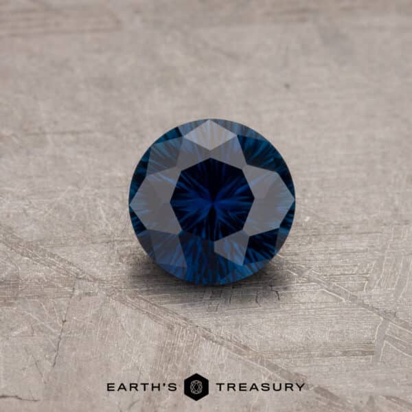 A close-up of a round brilliant-cut 1.53-Carat Royal Blue Ceylon Sapphire (Heated) resting on a textured surface. The gemstone has intricate facets, creating a striking star-like pattern when reflecting light. The text "EARTH'S TREASURY" is visible at the bottom of the image.