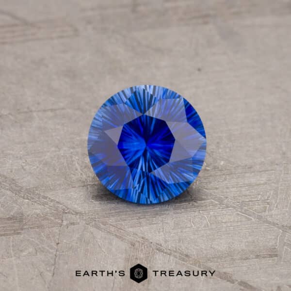 A close-up image features a 1.53-carat Royal Blue Ceylon Sapphire (Heated) with intricate facets, displayed on a textured gray surface. The gemstone boasts a vivid, rich blue hue and is elegantly presented by Earth's Treasury.