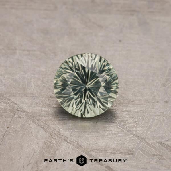 A 1.53-carat royal blue Ceylon sapphire, masterfully heated to enhance its brilliance, is displayed on a textured gray surface. The gem's intricate facets catch the light, creating a mesmerizing starburst pattern. The text "EARTH'S TREASURY" is visible below the gemstone.