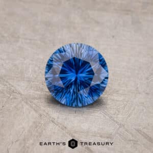 A 1.53-carat Royal Blue Ceylon Sapphire (heated), round and faceted, displayed on a textured grey surface. Beneath the gemstone, the logo and text "Earth's Treasury" are visible. The sapphire exhibits a deep blue hue with intricate, reflective facets.