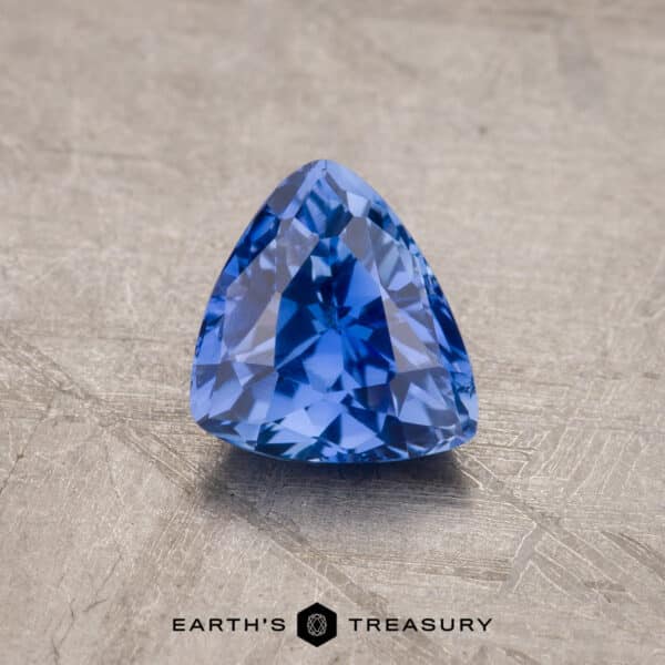 A vibrant royal blue, trillion-cut 1.53-carat Ceylon sapphire rests on a textured beige surface. The gem's facets catch the light, showcasing its clarity and depth of color. The photo features "Earth's Treasury" text at the bottom.