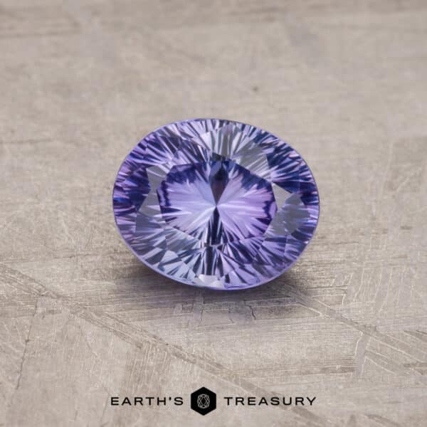 A 1.53-carat Royal Blue Ceylon Sapphire (heated), cut into an oval shape, is positioned on a textured gray surface. The gemstone's intricate facets produce a dazzling starburst pattern. The text "EARTH'S TREASURY" appears at the bottom of the image.