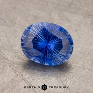 A close-up image of a faceted 1.53-carat Royal Blue Ceylon Sapphire with an intricate pattern, displayed on a textured gray surface. The sapphire is oval-shaped and reflects light, showcasing its vibrant color and precision cut. Text at the bottom reads "EARTH'S TREASURY.