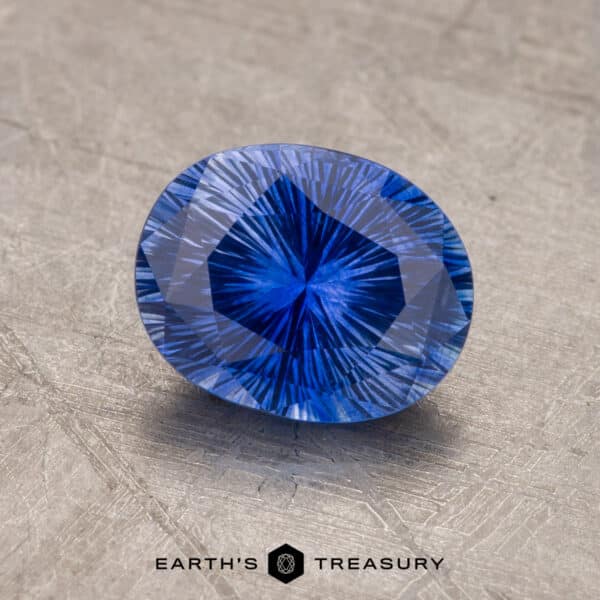 A close-up image of a faceted 1.53-carat Royal Blue Ceylon Sapphire with an intricate pattern, displayed on a textured gray surface. The sapphire is oval-shaped and reflects light, showcasing its vibrant color and precision cut. Text at the bottom reads "EARTH'S TREASURY.