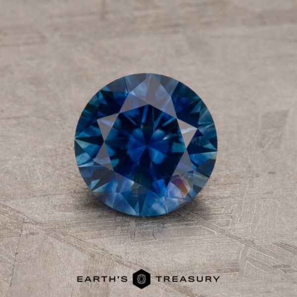 A round, brilliant-cut blue gemstone displayed against a textured background. The 1.53-carat Royal Blue Ceylon Sapphire (Heated) features intricate facets, showcasing deep blue hues and reflections of light. The bottom of the image includes the text "EARTH'S TREASURY" with a stylized logo.

