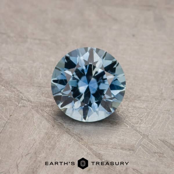 A close-up image of a faceted, round blue gemstone resting on a textured surface. The 1.53-Carat Royal Blue Ceylon Sapphire (Heated) is brilliantly cut, reflecting light and showcasing its internal facets. The words "EARTH'S TREASURY" are visible at the bottom of the image.