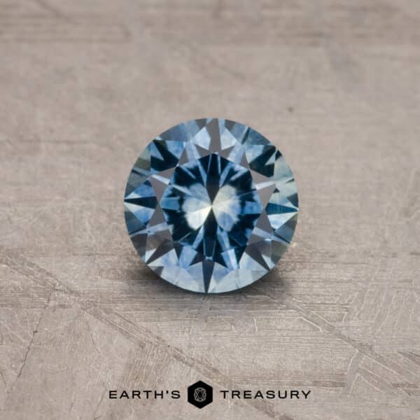 A brilliant round-cut 1.53-Carat Royal Blue Ceylon Sapphire (Heated) rests on a textured gray surface. The intricate facets of the stone catch and reflect light. At the bottom of the image, the logo "EARTH'S TREASURY" is visible.