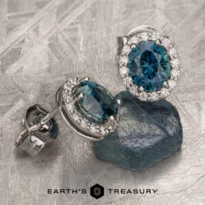 A pair of elegant earrings featuring oval-cut rich blue Montana sapphires, each surrounded by a halo of small sparkling diamonds. The earrings are set on a textured metallic surface along with a matching raw blue sapphire. The brand name "Earth's Treasury" is displayed at the bottom.