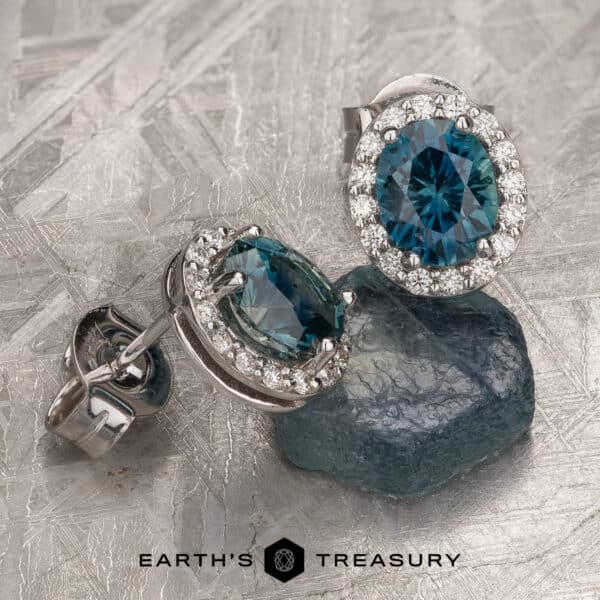 A pair of elegant earrings featuring oval-cut rich blue Montana sapphires, each surrounded by a halo of small sparkling diamonds. The earrings are set on a textured metallic surface along with a matching raw blue sapphire. The brand name "Earth's Treasury" is displayed at the bottom.