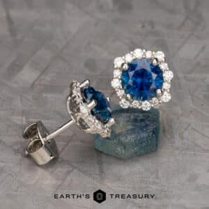 A pair of Montana Sapphire Halo Earrings featuring rich blue 7.0mm heated sapphires at the center, encircled by a halo of smaller white diamonds, set in silver. One earring is standing upright on a blue mineral, while the other is lying flat. Text at the bottom reads "Earth's Treasury.