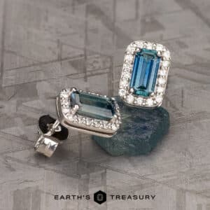 A pair of Montana Sapphire Halo Earrings featuring rich blue, 7.0mm heated stones surrounded by small clear gems. The earrings are resting on a textured gray surface, with the brand name "Earth's Treasury" visible at the bottom of the image.