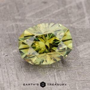 A 2.43-carat olive green demantoid garnet is displayed on a textured gray surface with the text "EARTH'S TREASURY" below.