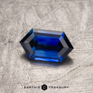 A hexagonal blue gemstone, a 1.05-carat Ceylon sapphire (heated), rests on a textured surface. The words "Earth's Treasury" are visible at the bottom of the image.