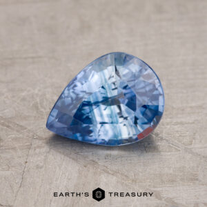 A 1.50-carat, pear-shaped, faceted Ceylon sapphire (heated) is placed on a textured surface with the text "Earth's Treasury" visible at the bottom of the image.