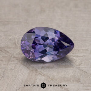 A 1.24-carat Madagascar sapphire (heated), featuring a teardrop shape and faceted cuts with shades of violet and blue, is displayed on a textured gray surface. The logo "EARTH'S TREASURY" is visible at the bottom.