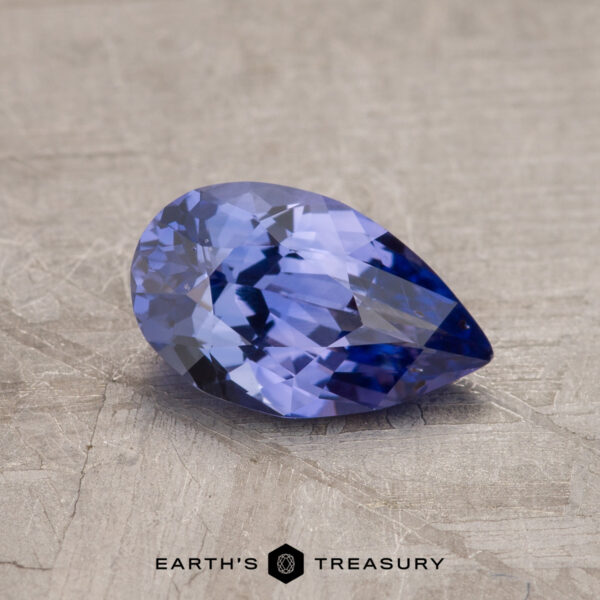 A pear-shaped, faceted blue gemstone rests on a textured surface. The text "EARTH'S TREASURY" is visible at the bottom of the image. This is the 1.15-Carat Ceylon Sapphire.